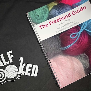 May include: A black tote bag with the text "HALF HOOKED" and a yarn ball graphic. A spiral-bound book titled "The Freehand Guide" by Casey Ruiz is visible, with colorful yarn in the background.