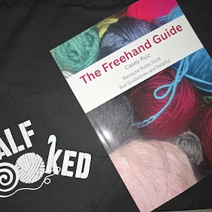 May include: A black tote bag with the text "HALF HOOKED" and a yarn ball graphic. Beside it is a book titled "The Freehand Guide" by Casey Ruiz, with colorful yarn skeins in the background. The book's subtitle reads "Because Rules Suck But Guidelines are Helpful."