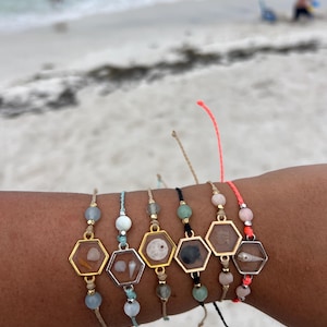 May include: A collection of six handmade bracelets, each featuring a hexagonal charm with a seashell or sea-themed element encased within. The bracelets have various colored beads and cords, with gold and silver-toned metal accents. The background is a sandy beach with ocean waves.