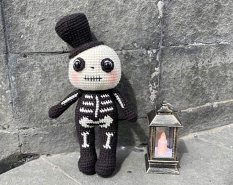 Puede incluir: Muñeca de esqueleto de ganchillo hecha a mano con un sombrero de copa negro. La muñeca tiene una cabeza blanca con ojos negros y una boca cosida. El cuerpo del esqueleto es negro con detalles óseos blancos. Una pequeña linterna dorada con una vela falsa está cerca.