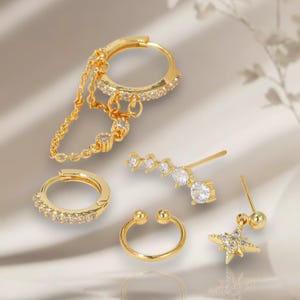 Op de afbeelding: Een verzameling goudkleurige oorbellen, waaronder creolen, een creool met ketting, een ster-vormige oorbel, een gebogen oormanchet en een oor-climber. Elk stuk is versierd met heldere, sprankelende edelstenen, wat een elegante set creëert.