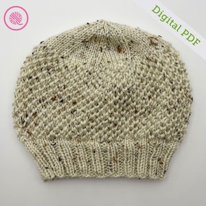 Puede incluir: Un gorro de punto color crema con motas marrones y negras. El gorro tiene un borde acanalado y un patrón de puntada texturizada. Las palabras "Digital PDF" están impresas en verde. Un logotipo de hilo rosa y blanco está en la esquina superior izquierda.