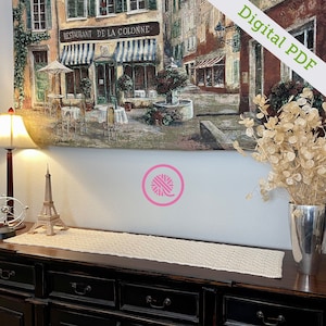May include: A crocheted table runner in a cream color with a textured pattern. The runner is displayed on a dark wood table with drawers. A painting of a Parisian street scene hangs on the wall behind the table. A small Eiffel Tower figurine and a lamp are on the table.