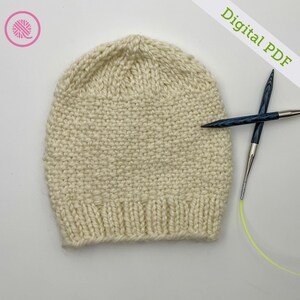 Puede incluir: Gorro tejido de color crema con borde acanalado. El gorro está hecho de hilo grueso y tiene una apariencia texturizada. Las agujas de tejer se colocan junto al gorro. Las palabras "Digital PDF" están escritas en verde.