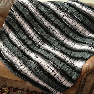 Puede incluir: Una manta de punto a rayas en tonos de negro, gris y rosa. La manta está colocada sobre un sofá de cuero marrón.