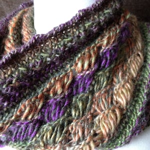 May include: A hand-knitted cowl in shades of purple, green, brown, and beige. The cowl is made with a chunky yarn and has a textured stitch pattern.