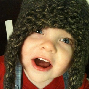Può includere: Un bambino piccolo che indossa un cappello di lana marrone e nero con una camicia a quadri blu e bianca sotto. Il bambino sorride e ha gli occhi azzurri.