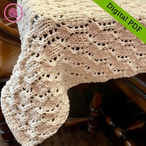 Puede incluir: Primer plano de un camino de mesa de ganchillo con un patrón de diamante en color crema. El camino de mesa está drapeado sobre una mesa de madera.