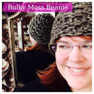 May include: A woman wearing a brown and black knit beanie with a bulky moss stitch pattern. She is wearing glasses and smiling.