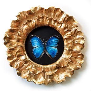 Puede incluir: Una mariposa azul con venas negras está enmarcada en un marco dorado en forma de flor. La mariposa está centrada en el marco y tiene las alas extendidas.