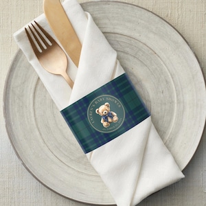 May include: A white cloth napkin with a gold fork and wooden knife, wrapped and held together by a dark green and blue plaid band. The band features a teddy bear illustration and the text "LAUREN'S BABY SHOWER OCTOBER 15, 2021".