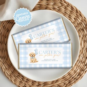Op de afbeelding: Twee chocoladereepverpakkingen met een blauw geruit patroon en gouden accenten. Elke verpakking heeft een cartoon puppy, de tekst "CARTER'S one golden year!" en "THANK YOU FOR CELEBRATING WITH US!" Een "Printable Canva Template" logo staat linksboven.