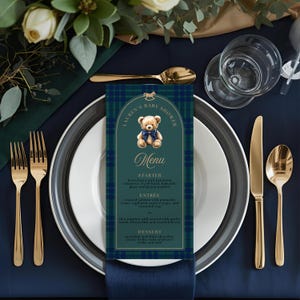 May include: A baby shower menu card featuring a teddy bear illustration, set against a navy blue and green plaid pattern. The card is on a white plate with gold cutlery and a gold spoon. The menu includes the text "Lauren's Baby Shower" and the word "Menu".