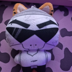May include: A plush toy featuring a white, stylized character head with large black sunglasses, prominent eyebrows, and orange-tipped ears. The character has a white torso with black pants and a small metal keychain attachment at the top of the head.