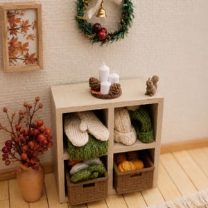 Könnte beinhalten: Ein Miniatur-Holzregal mit gestrickten Fäustlingen, Mützen und geflochtenen Körben voller kleiner Kürbisse. Auf der Oberseite stehen drei weiße Stumpenkerzen, Tannenzapfen und eine Eichhörnchenfigur, ergänzt durch einen Kranz und gerahmte Kunst.