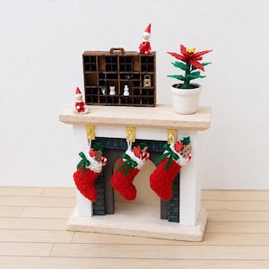 Op de afbeelding: Een miniatuur witte open haard met een licht houten schoorsteenmantel, drie rode gebreide kousen met groene linten, een bruine houten vakkenkast met twee kleine elfenbeeldjes en een rode kerstster in een witte pot.