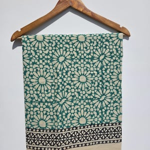 May include: A patterned fabric in shades of green and cream, displayed on a wooden hanger. The fabric showcases a repeating floral motif, with a border of black and cream geometric patterns. Likely a scarf or textile.