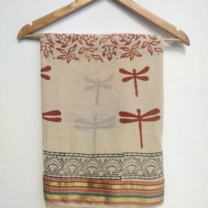 May include: A beige textile with a dragonfly pattern and floral border, hanging on a wooden hanger. The dragonfly design is in a deep red colour, with a decorative border in black, gold, and red. The textile is likely a scarf or shawl.