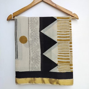 May include: A rectangular scarf with a cream background and black, gold, and white geometric patterns. The scarf features a black border with a gold trim, a black triangle design, and a floral pattern.