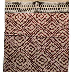 May include: A folded textile with a cream-colored top and a geometric pattern in burgundy and cream. The pattern consists of diamond shapes and a black border with a repeating design.