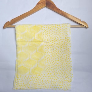 May include: A yellow and white scarf with a repeating pattern of yellow fan-like shapes and yellow polka dots. The scarf is hanging on a wooden hanger against a white background.
