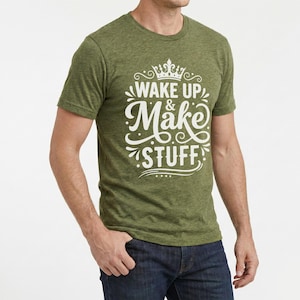 以下が含まれることがあります： 白い文字で「WAKE UP & Make STUFF」と書かれた緑色のクルーネックTシャツ。シャツには単語の上に王冠のデザインがあります。モデルはブルージーンズを着用しています。
