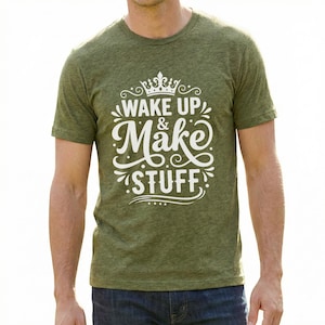 以下が含まれることがあります： 「WAKE UP & Make STUFF」の白文字がプリントされたグリーンのTシャツ。デザインには王冠と装飾的な要素が含まれています。半袖のTシャツは、質感のある素材で作られています。