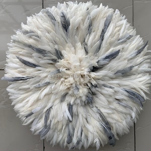 May include: Circular wall hanging composed of white and gray feathers. The feathers are arranged in a radial pattern, creating a textured, layered effect. The center is a lighter shade of white, with darker gray feathers interspersed throughout.