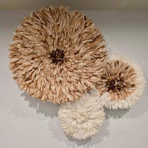 Peut inclure: Trois décorations murales circulaires en plumes. La plus grande est de couleur marron clair, la seconde est blanche et marron, et la plus petite est blanche. Elles sont montées sur un mur gris clair, créant un effet texturé et naturel.