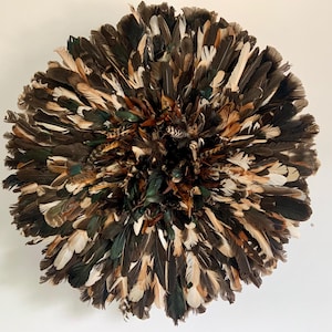 Op de afbeelding: Een ronde wanddecoratie gemaakt van veren in tinten bruin, zwart, groen en crème. De veren zijn in een cirkelvormig patroon gerangschikt, waardoor een textuur en gelaagd effect ontstaat. Dit decoratieve stuk voegt een natuurlijke en aardse toets toe.