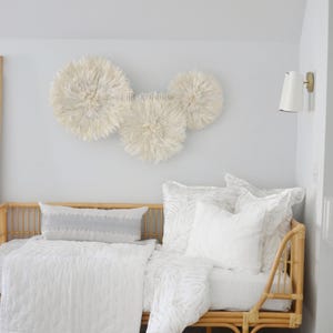 Peut inclure: Trois décorations murales en plumes de couleur crème au-dessus d'un lit avec une literie et des oreillers blancs. Le cadre de lit est en osier brun clair. Une applique murale est fixée au mur.