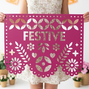 May include: A magenta paper banner with the word "FESTIVE" cut out in white. The banner features decorative cutouts of flowers, leaves, hearts, and geometric shapes. The banner is held up by a person.