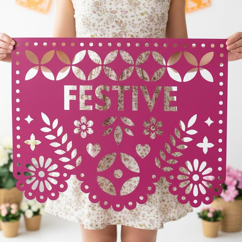 May include: A magenta paper banner with the word "FESTIVE" cut out in white. The banner features decorative cutouts of flowers, leaves, hearts, and geometric shapes. The banner is held up by a person.