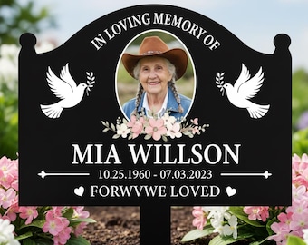 Op de afbeelding: Een zwarte herdenkings-tuinsteker met de tekst IN LOVING MEMORY OF, een ronde foto van een persoon met een cowboyhoed, twee witte duiven, roze bloemaccenten, de naam MIA WILLSON, de data 10.25.1960 - 07.03.2023 en FORWVWE LOVED.