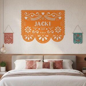 May include: An orange papel picado banner with the name "JACKI" in white, surrounded by floral and heart cutouts. Two smaller, decorative banners hang on either side. The scene is set above a bed with decorative pillows.