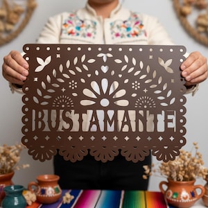 May include: A brown, laser-cut decorative sign with the name "BUSTAMANTE" in a decorative font. The sign features intricate floral and geometric cut-out designs along the edges and within the letters. The sign is held up by a person.