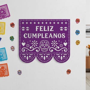 May include: A purple banner with the words "FELIZ CUMPLEANOS" in white, decorated with skull and floral designs. The banner is surrounded by colorful skull decorations and papel picado banners, perfect for a Day of the Dead celebration.
