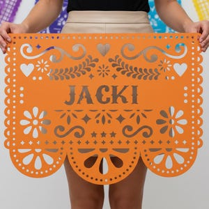 May include: An orange paper banner with the name "JACKI" cut out in the center. The banner features decorative cut-out designs of hearts, flowers, and stars along the edges and top.