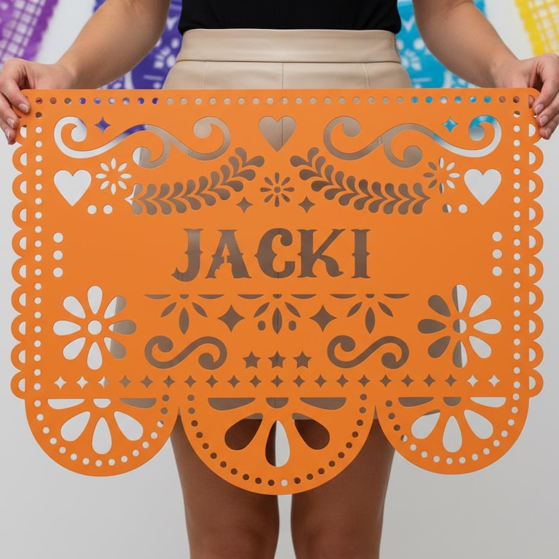 May include: An orange paper banner with the name "JACKI" cut out in the center. The banner features decorative cut-out designs of hearts, flowers, and stars along the edges and top.