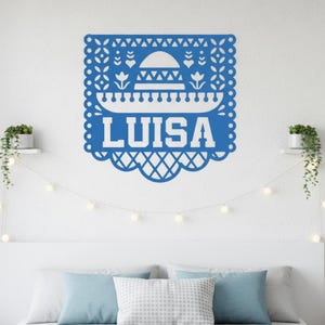 May include: A blue paper cut-out decoration with the name "LUISA" in white. The design includes a sombrero, floral and geometric patterns, and a scalloped edge. The decoration is mounted on a white wall.
