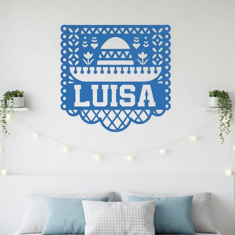 May include: A blue paper cut-out decoration with the name "LUISA" in white. The design includes a sombrero, floral and geometric patterns, and a scalloped edge. The decoration is mounted on a white wall.