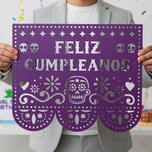 May include: A purple paper banner with the words "FELIZ CUMPLEANOS" cut out in silver. The banner features decorative cutouts of skulls, flowers, hearts, and geometric shapes. The banner is held by a person.