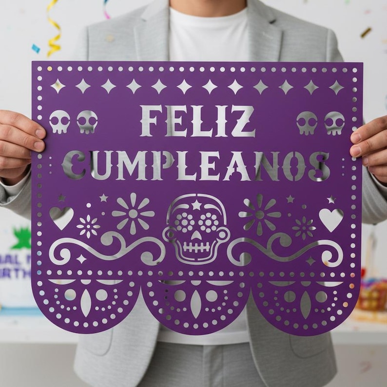 May include: A purple paper banner with the words "FELIZ CUMPLEANOS" cut out in silver. The banner features decorative cutouts of skulls, flowers, hearts, and geometric shapes. The banner is held by a person.