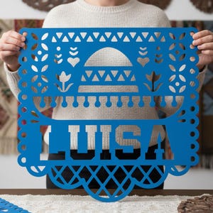 May include: A vibrant blue papel picado banner with the name "LUISA" cut out in bold letters. The decorative banner features intricate designs, including hearts, flowers, and geometric patterns, with a scalloped edge.