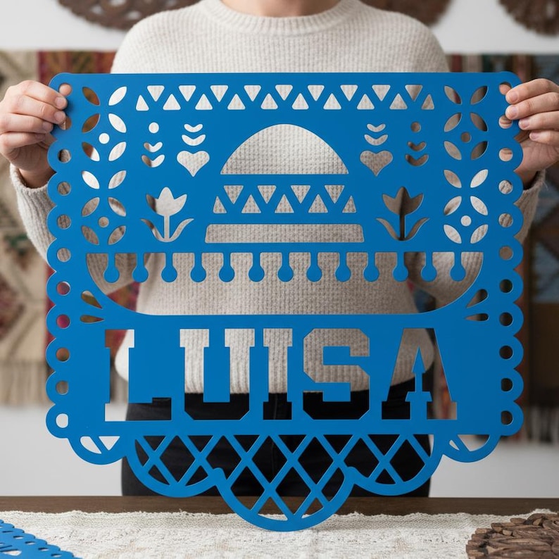 May include: A vibrant blue papel picado banner with the name "LUISA" cut out in bold letters. The decorative banner features intricate designs, including hearts, flowers, and geometric patterns, with a scalloped edge.