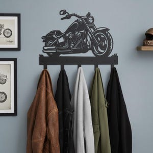 May include: A black metal motorcycle-shaped coat rack with five hooks. The motorcycle outline is above the hooks, and various jackets and coats in brown, black, grey, and green hang from the hooks. Two framed motorcycle prints are on the wall.