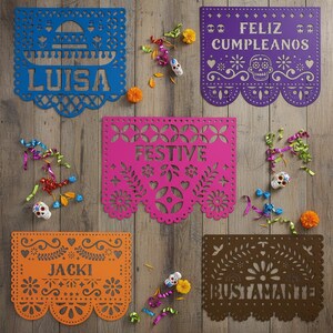 May include: A collection of colorful papel picado banners with intricate cut-out designs. The banners are in blue, purple, pink, orange, and brown, with names and festive phrases. Decorative skulls and colorful confetti are scattered around the banners.