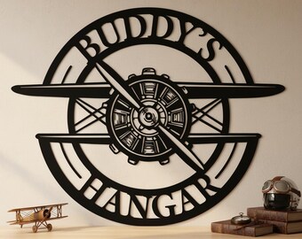 May include: A circular black metal wall sign featuring a stylized airplane propeller and engine design. The text BUDDY'S is cut out along the top arc, and the word HANGAR is cut out along the bottom arc of the circular frame structure.