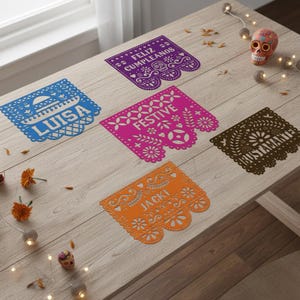May include: A wooden table displays colorful papel picado banners with cut-out designs and text. The banners are blue, purple, pink, brown, and orange. Text includes "LUISA", "FELIZ CUMPLEANOS", "FESTIVE", "BUSTAMANTE", and "JACKI". A decorative skull and string lights are also present.