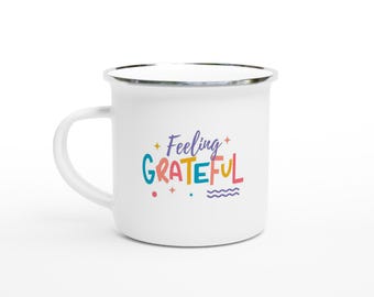 May include: White enamel mug with a silver rim and handle. The mug features the words "Feeling Grateful" in colorful, playful lettering. The design includes stars, dots, and a wavy line, creating a cheerful and positive vibe.